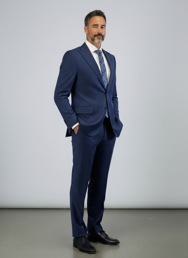 A man with gray hair and a beard stands against a plain background, wearing the FUBU Indigo Solid Peak Lapel 2-Piece Suit by Fubu The Colle—a slim fit ensemble featuring a peak lapel jacket, paired with a white shirt and patterned tie, his hands casually in his pockets.