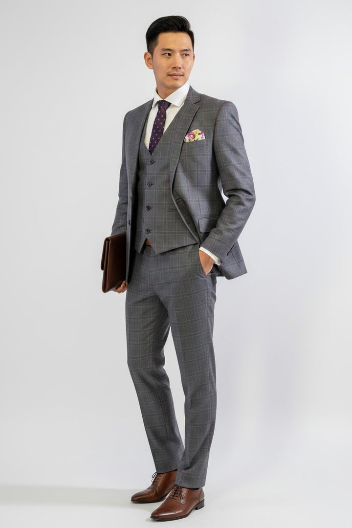 A man is wearing the FUBU Charcoal with Purple Windowpane Vested Suit by Fubu The Colle, paired with a white shirt, purple patterned tie, and pocket square. The suit features a stretch armhole design for added comfort. He holds a brown briefcase and stands against a plain white background.