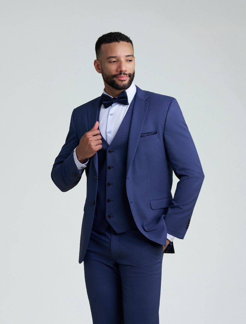 A man wearing the Allure Men's Royal Blue Tailored Fit Suit Jacket, styled with a white dress shirt and black bow tie, stands confidently—one hand adjusting his jacket, the other in his pocket.