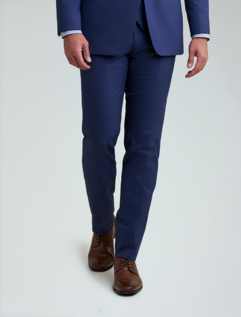 A man wears Allure Men's Royal Blue Tailored Fit Dress Pants, paired with a coordinating suit and brown dress shoes, standing against a plain light background.