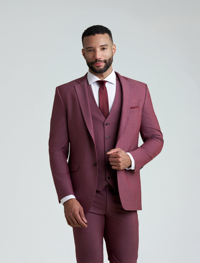A man stands against a plain light background, wearing the Allure Men's Burgundy Tailored Fit Suit Jacket styled with a matching vest, white dress shirt, and burgundy tie.