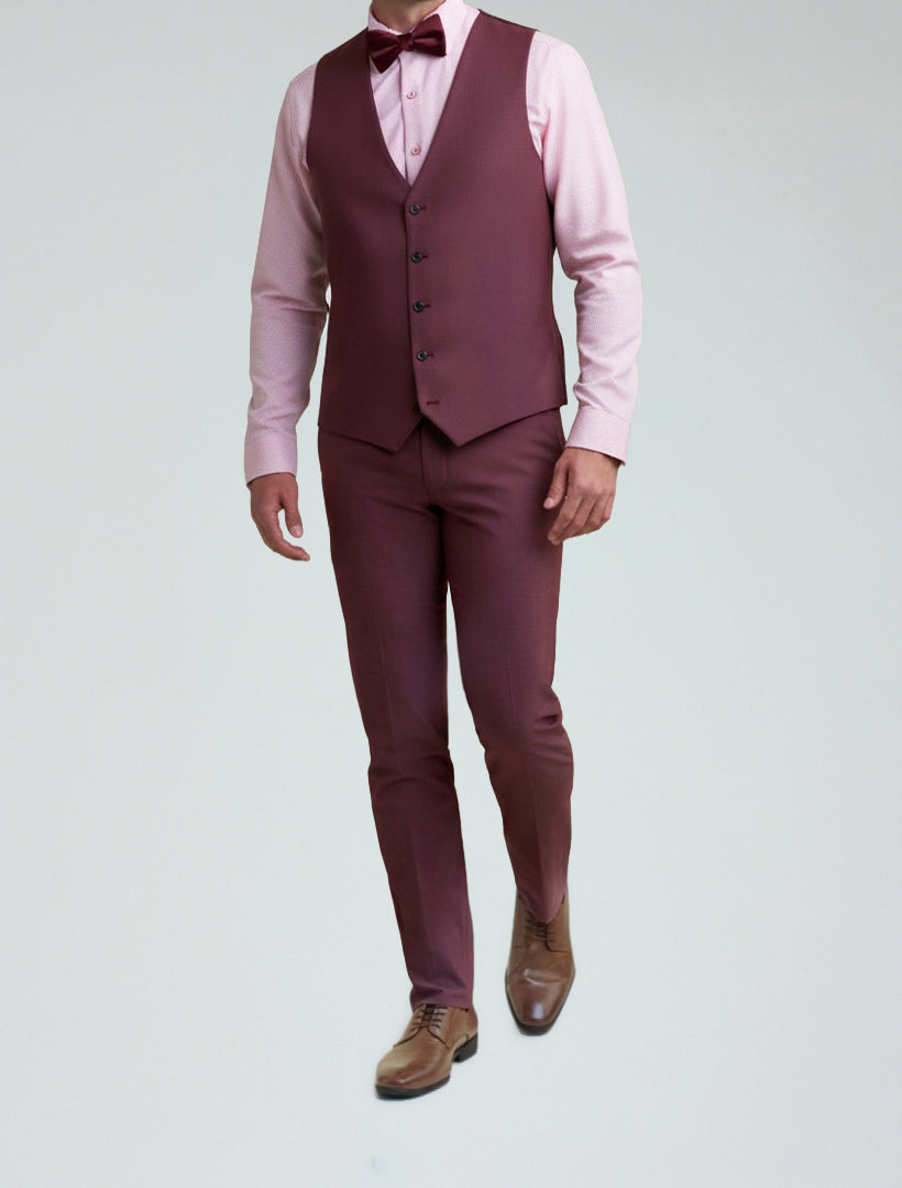 A man wearing the Allure Men's Burgundy Tailored Fit Vest, styled with a matching jacket, pink dress shirt, bow tie, and brown dress shoes, stands against a light gray background.