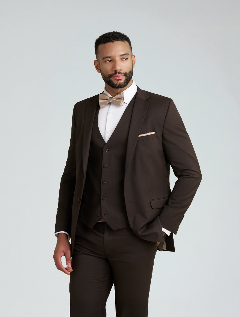 A man stands slightly sideways against a plain background, wearing the Allure Men's Brown Tailored Fit Suit Jacket with notch lapels, a matching vest, light beige bow tie, and pocket square.