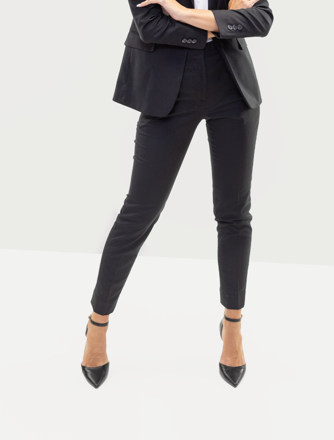 A person wearing a Couture 1910 Women's Black Suit Separates Pants and heels, standing with arms crossed, radiates confidence with their sleek fit and tailored design.