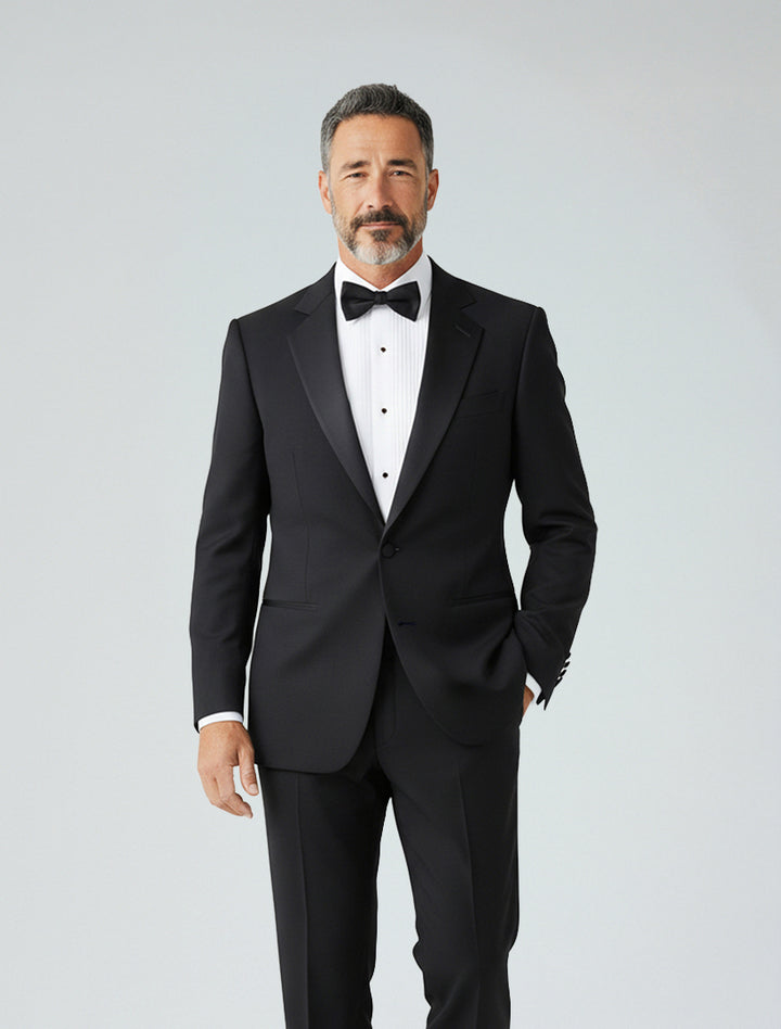 Man wears the FUBU Classic Notch Lapel Black Tuxedo by Fubu The Colle, styled with a white dress shirt and black bow tie, standing against a plain light background.