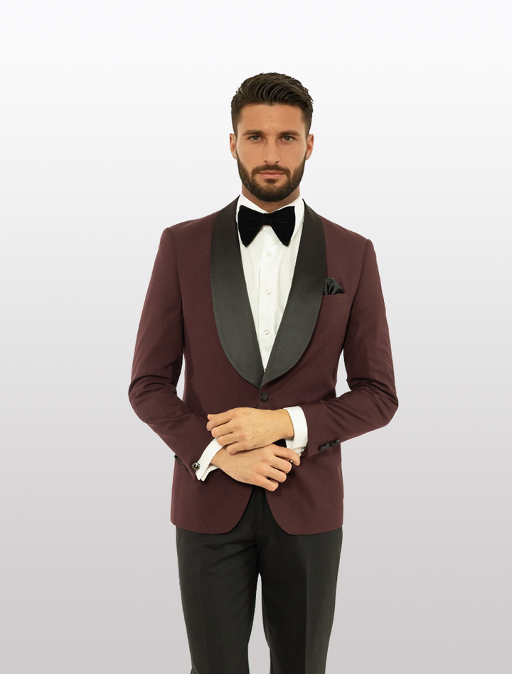 A man wears the FUBU Burgundy Shawl Lapel Slim Fit Tuxedo by Fubu The Colle, paired with a white shirt, black bow tie, and black pants, standing against a plain light background.