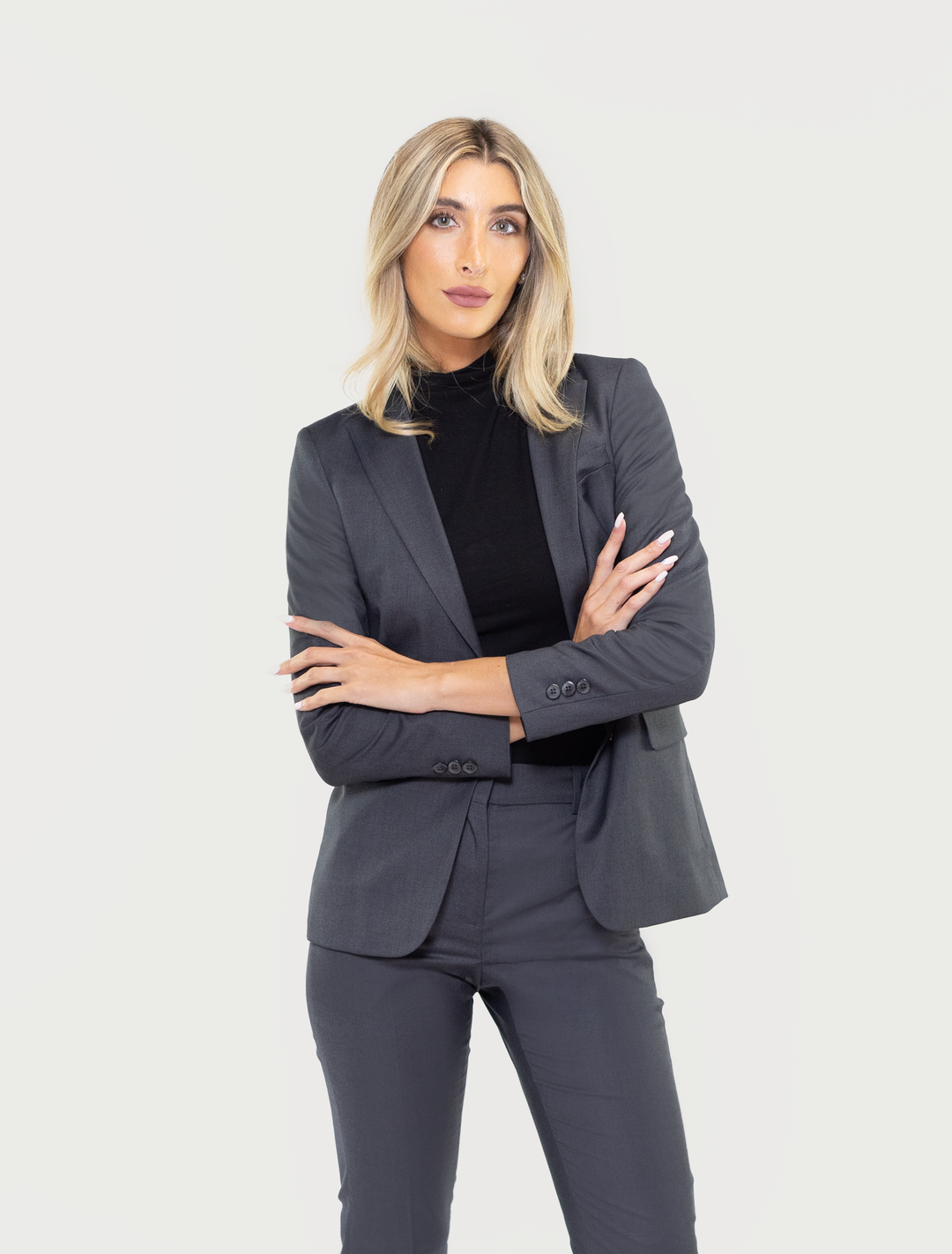 A woman in a Couture 1910 charcoal grey suit separates jacket and a black turtleneck stands with arms crossed against a white background.