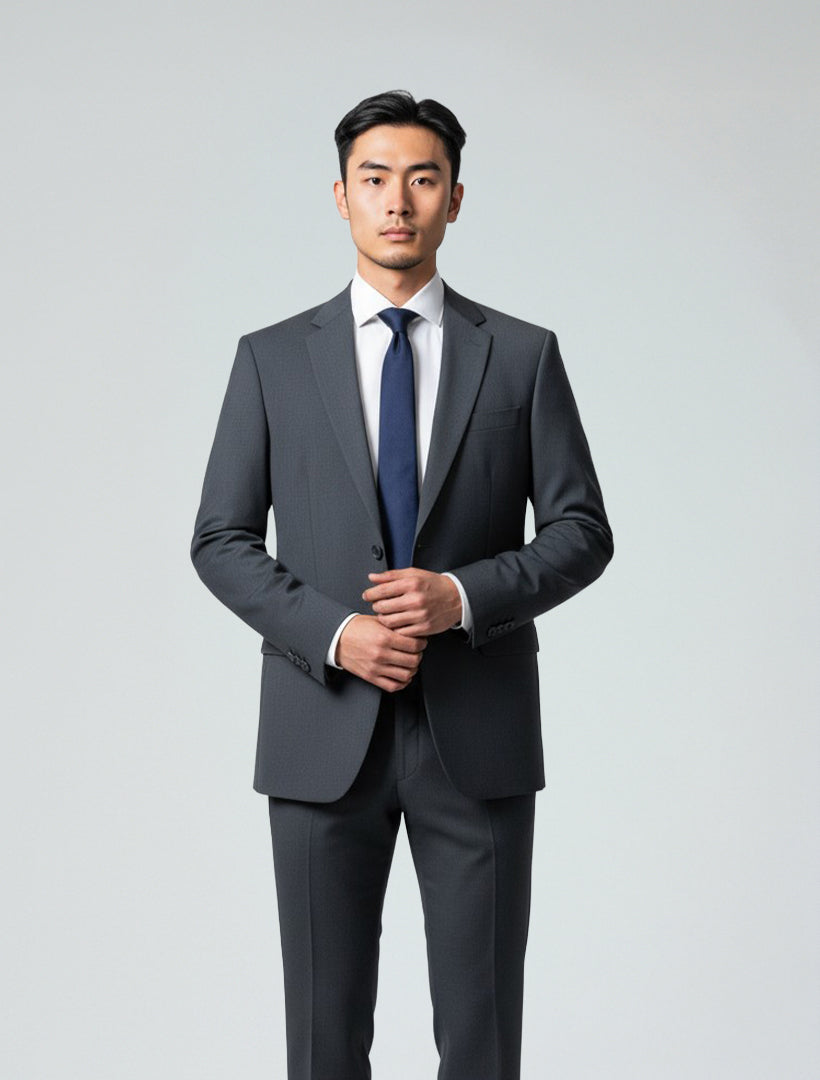 A man wears the Karako Men Mens Charcoal Suit with a white shirt and navy tie, standing against a plain light background—a stylish option for wedding attire.