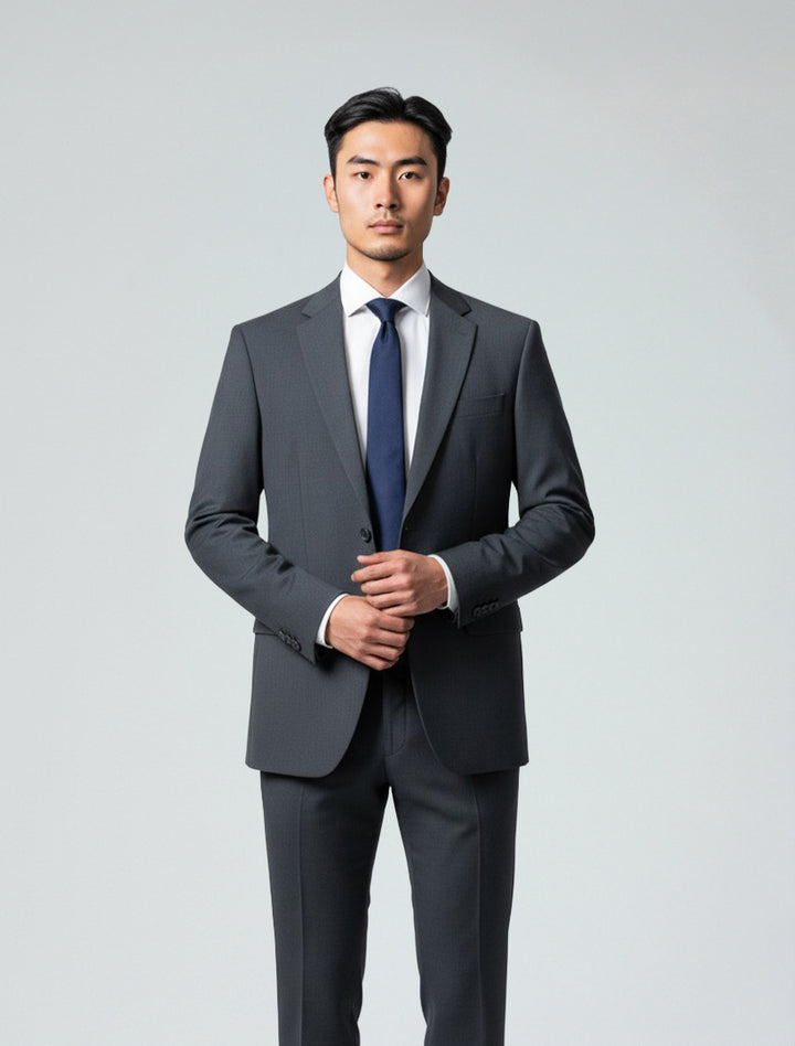 A man wears the Karako Men Mens Charcoal Suit with a white shirt and navy tie, standing against a plain light background—a stylish option for wedding attire.