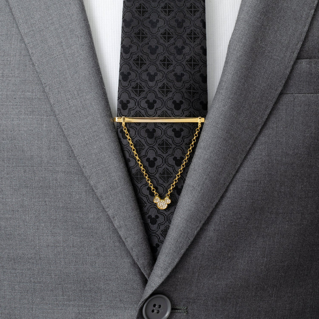 A Disney Mickey Mouse Gold Crystal Chain Tie Bar with a dangling chain attached to a dark grey patterned tie worn with a grey suit.