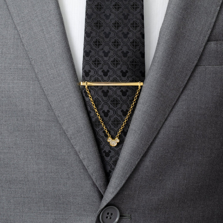 A Disney Mickey Mouse Gold Crystal Chain Tie Bar with a dangling chain attached to a dark grey patterned tie worn with a grey suit.