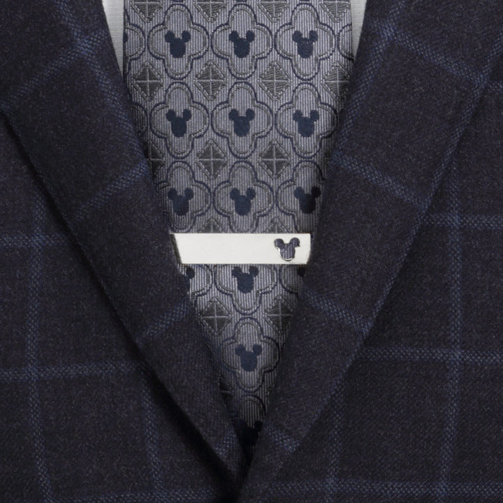 A silver-toned Disney Mickey Mouse Cutout Tie Bar featuring a small cutout, worn over a patterned gray tie and a dark plaid suit jacket.
