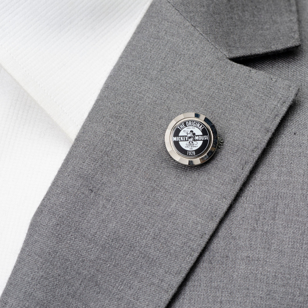 A round Disney Mickey Mouse Original Lapel Pin labeled “The Original Mickey Mouse 1928” attached to a gray suit lapel.