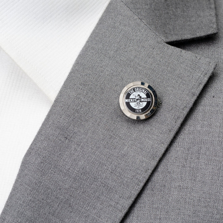 A round Disney Mickey Mouse Original Lapel Pin labeled “The Original Mickey Mouse 1928” attached to a gray suit lapel.