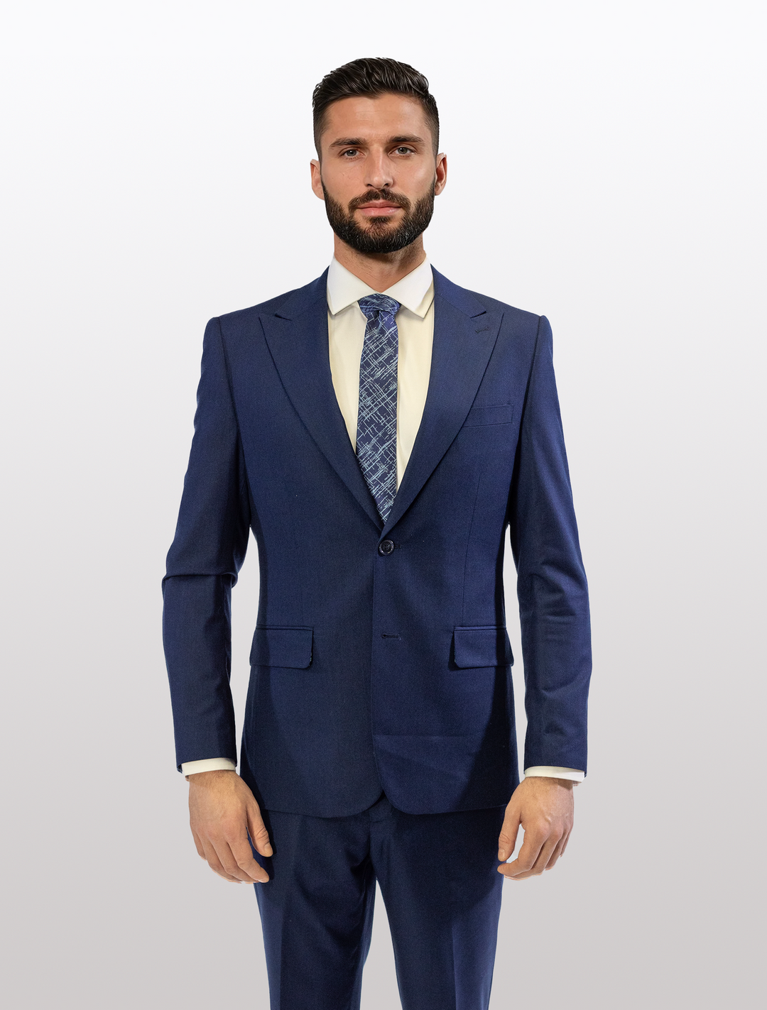 A man stands confidently against a plain white background wearing the FUBU Indigo Solid Peak Lapel 2-Piece Suit, by Fubu The Colle, complemented by a patterned tie and white shirt.