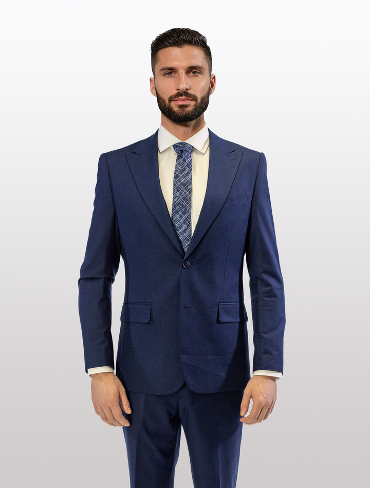 A man stands confidently against a plain white background wearing the FUBU Indigo Solid Peak Lapel 2-Piece Suit, by Fubu The Colle, complemented by a patterned tie and white shirt.