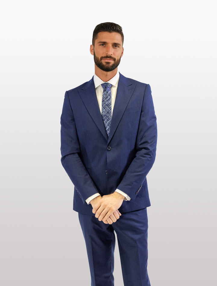 A man models the FUBU Indigo Solid Peak Lapel 2-Piece Suit from Fubu The Colle, complete with a tie, against a plain white background, highlighting the elegance of its stretch armholes.