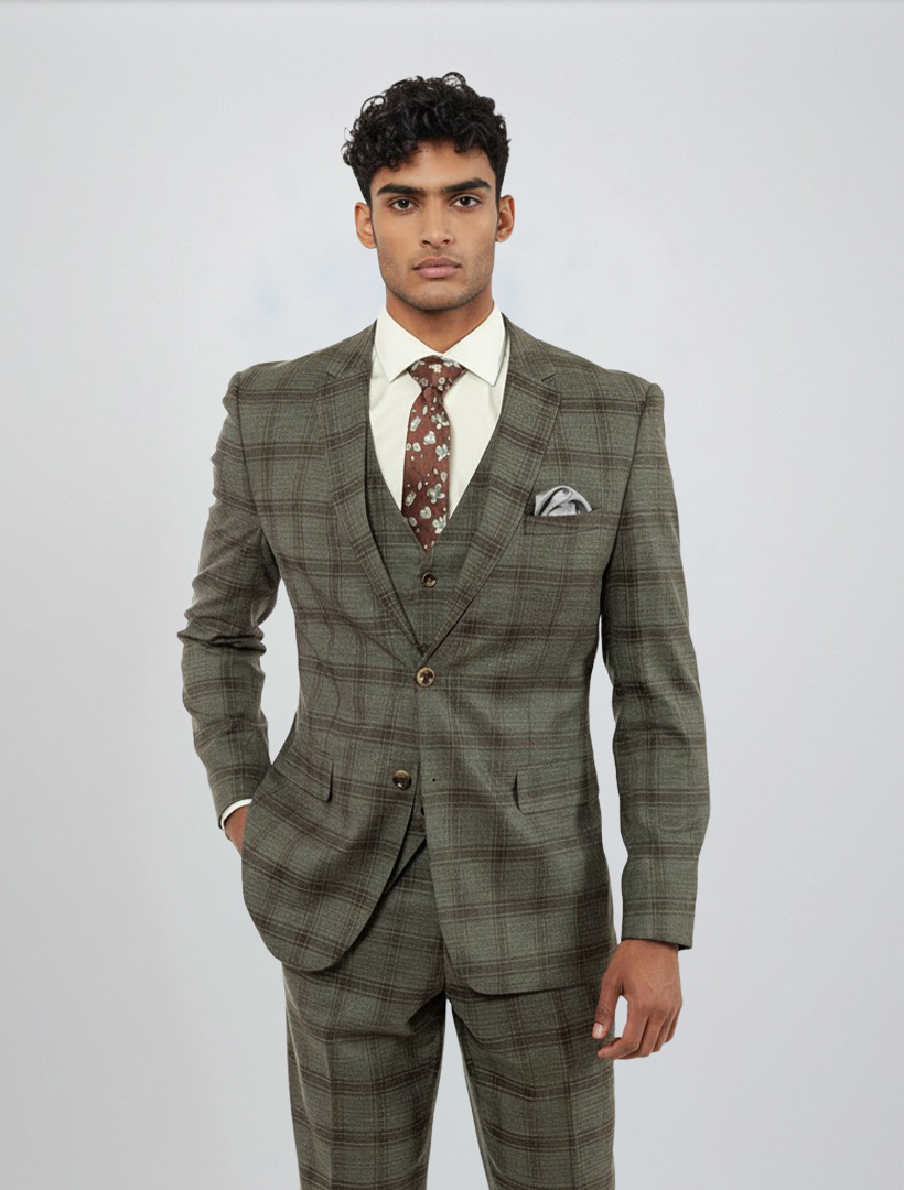 A man stands facing forward against a plain light background, wearing the FUBU Charcoal with Brown Plaid Vested Suit by Fubu The Colle. The slim fit suit, paired with a white shirt, patterned brown tie, and pocket square, features stretch armholes for added comfort and mobility.