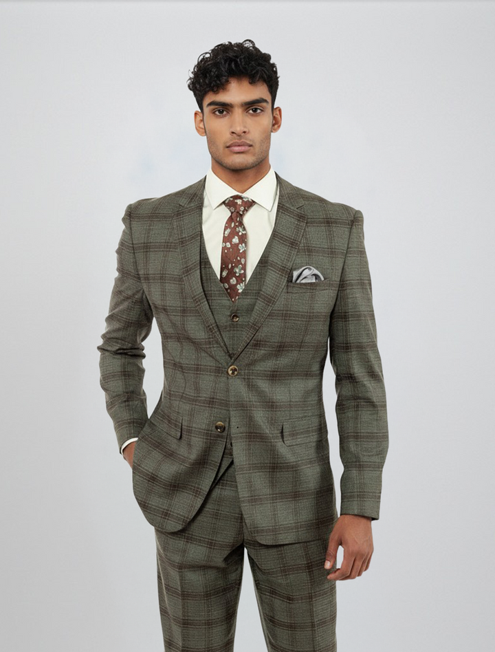 A man stands facing forward against a plain light background, wearing the FUBU Charcoal with Brown Plaid Vested Suit by Fubu The Colle. The slim fit suit, paired with a white shirt, patterned brown tie, and pocket square, features stretch armholes for added comfort and mobility.