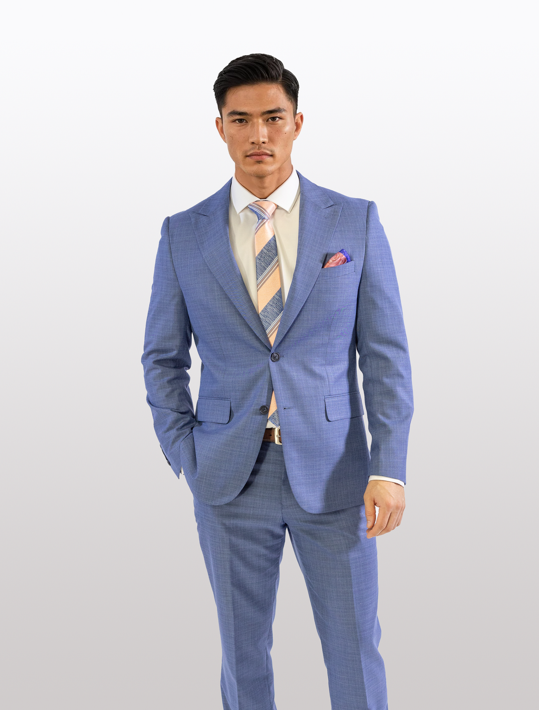 Wearing the FUBU Stone Blue Peak Lapel 2-Piece Suit by Fubu The Colle, a man stands against a plain white background. He pairs it with a peach and white tie, displays its sleek stretch armhole design, and keeps his left hand in his pocket while maintaining a neutral expression.