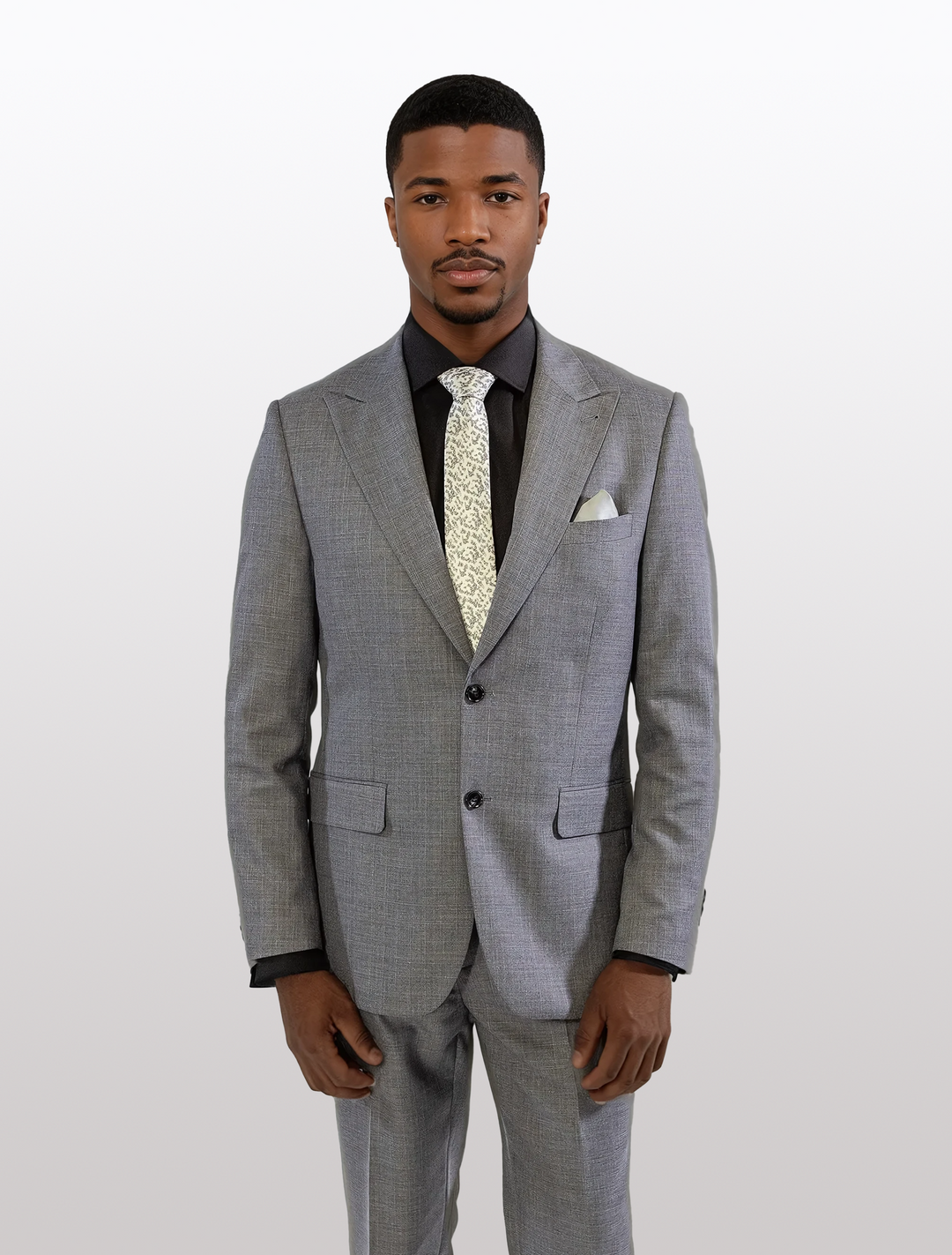 A man wearing the FUBU Iron Grey Peak Lapel 2-Piece Suit by Fubu The Colle, matched with a black shirt and patterned tie, stands against a plain white background.