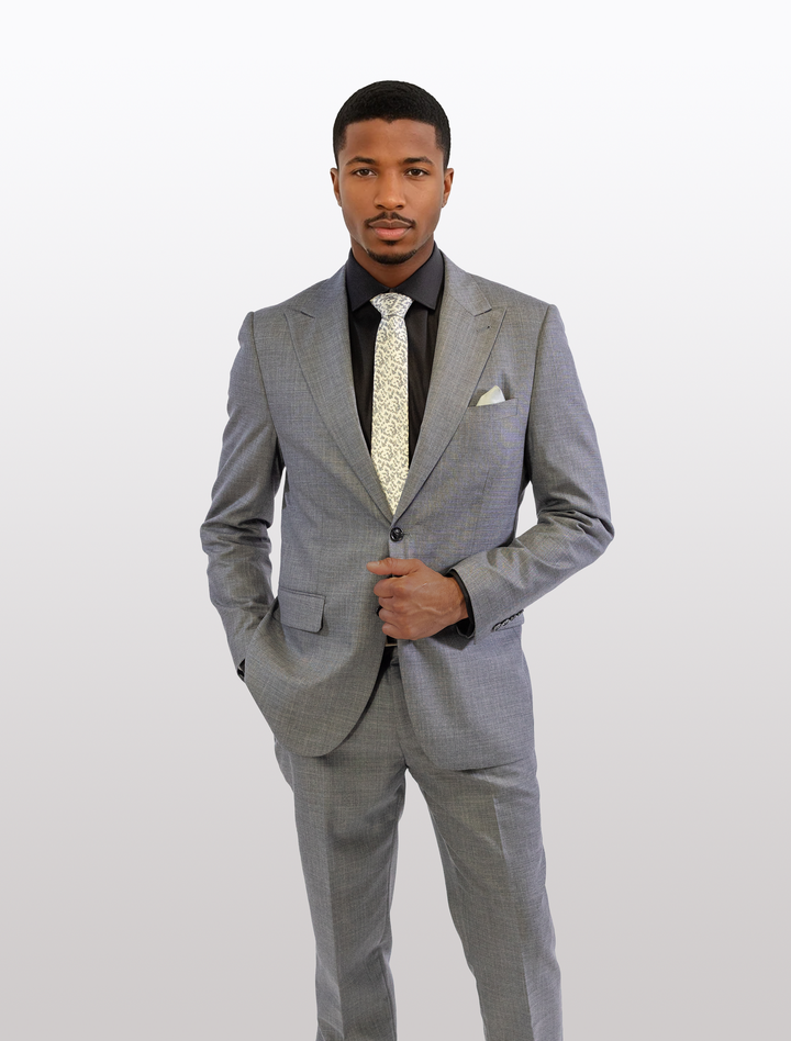 A man wearing the FUBU Iron Grey Peak Lapel 2-Piece Suit by Fubu The Colle, with a patterned tie, stands against a plain background, hands in pockets, looking forward.