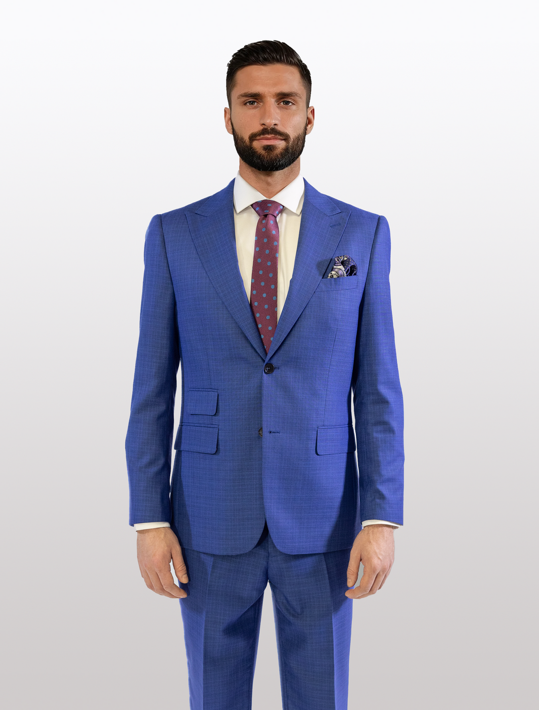 Man in a FUBU Cobalt Blue Peak Lapel 2-Piece Suit by Fubu The Colle, wearing a white shirt and a polka dot tie, standing against a plain background.