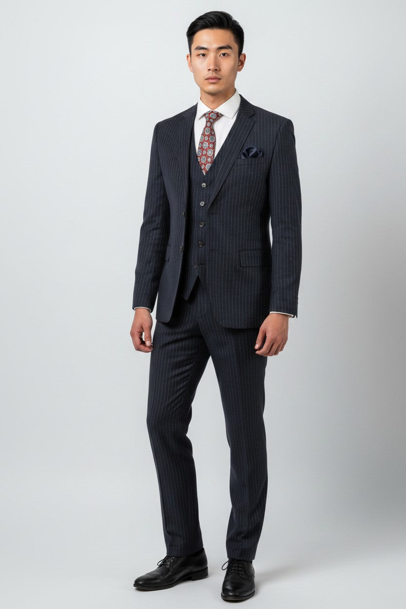 A man stands against a plain background wearing the FUBU Navy with White Pinstripe Vested Suit by Fubu The Colle, featuring a modern fit with stretch armholes, paired with a white dress shirt, patterned tie, vest, and black dress shoes.