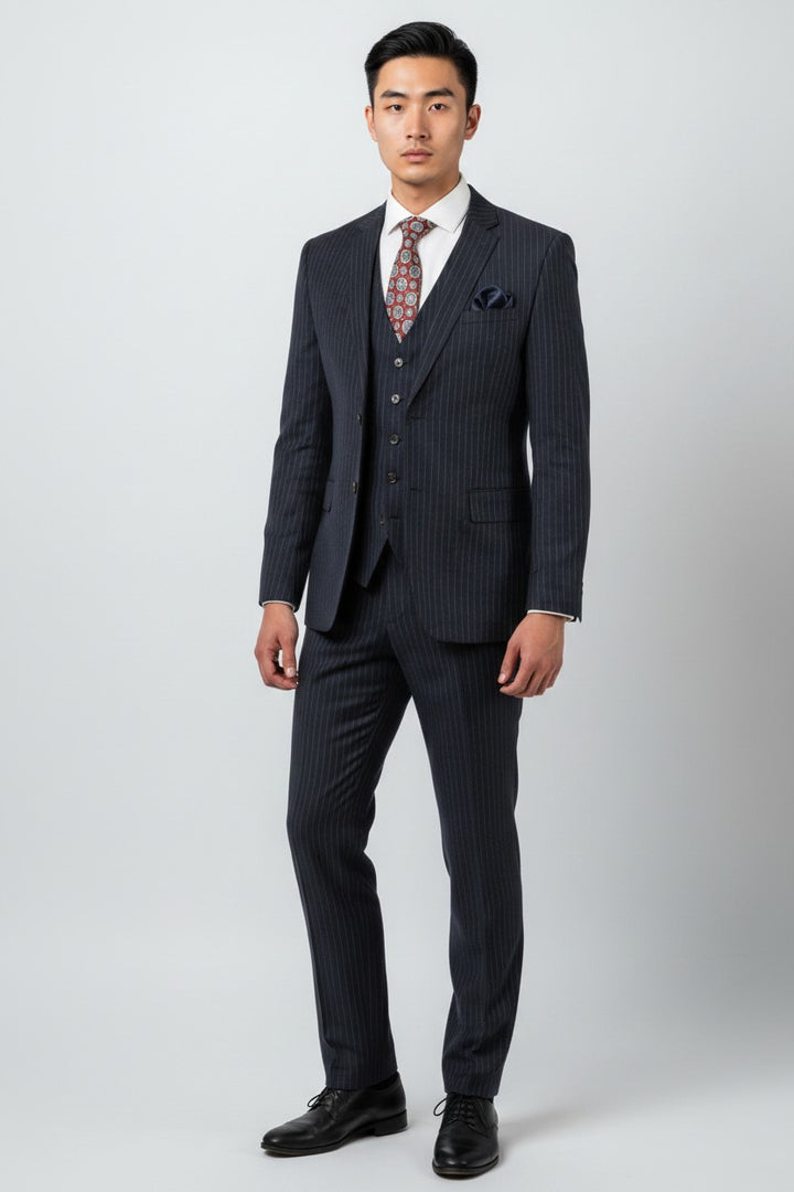 A man stands against a plain background wearing the FUBU Navy with White Pinstripe Vested Suit by Fubu The Colle, featuring a modern fit with stretch armholes, paired with a white dress shirt, patterned tie, vest, and black dress shoes.