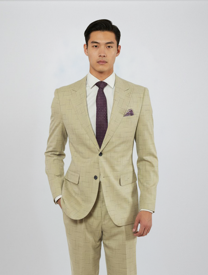 A man wearing the FUBU Tan with Navy Fading Windowpane Peak Lapel 2-Piece Suit by Fubu The Colle, paired with a white dress shirt and dark patterned tie, stands against a plain light background.