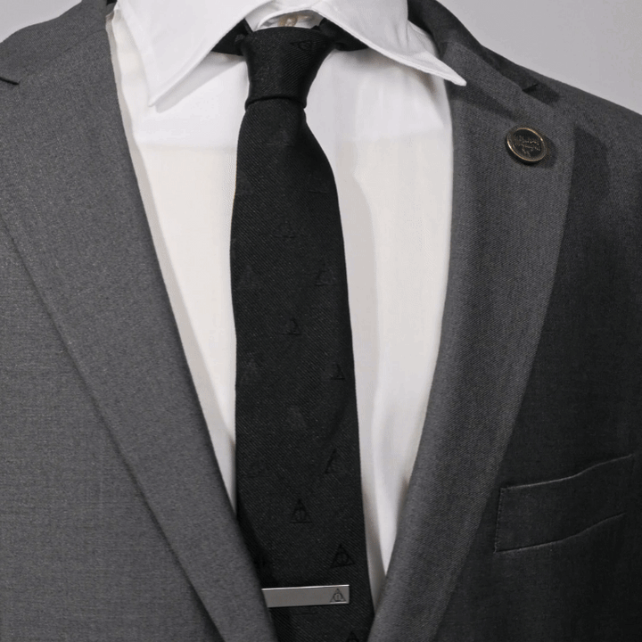 A close-up of a person in a grey suit and white shirt, wearing a Harry Potter tie patterned with Deathly Hallows symbols and secured with a Harry Potter Deathly Hallows “Always” Hidden Message Tie Bar.