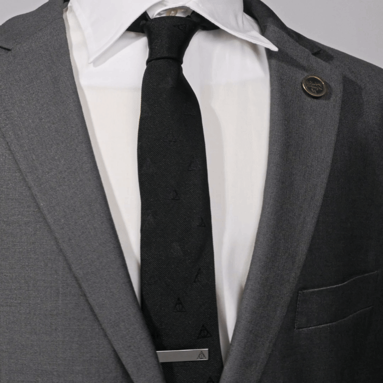 A black silk Cufflinks.com Harry Potter Deathly Hallows Tie with a repeating symbol pattern, worn with a white dress shirt, gray suit jacket, and a metallic tie clip.