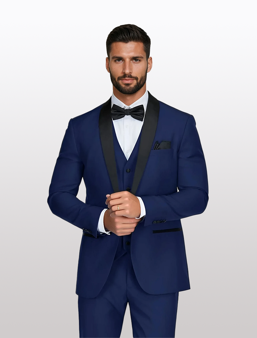 A man in a tailored-fit Men's Indigo Shawl Lapel Tuxedo by Gino Vitale, featuring black lapels and a bow tie, stands against a plain background adjusting his cufflinks. The Tetron Rayon fabric of the ensemble provides a polished finish.