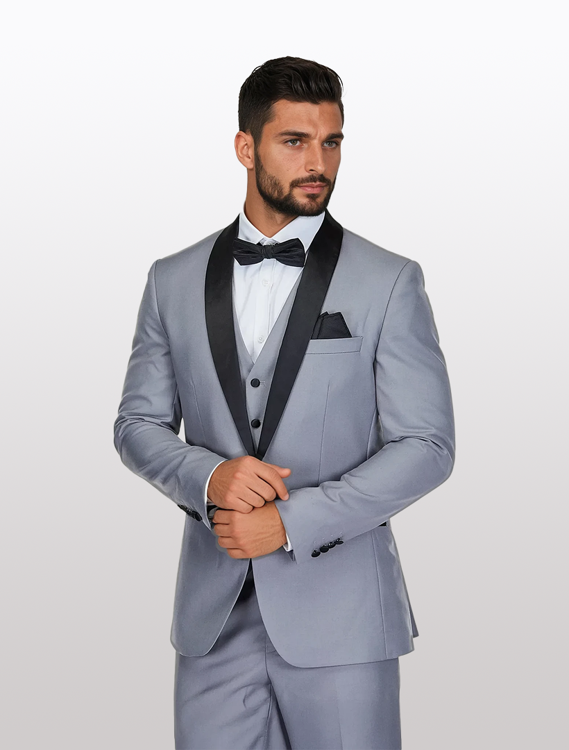 A man in a Gino Vitale Men's Light Grey Shawl Lapel Tailored Fit Tuxedo, featuring Tetron Rayon fabric, wears a black bow tie. He stands with hands clasped, gazing to the side against a plain backdrop.