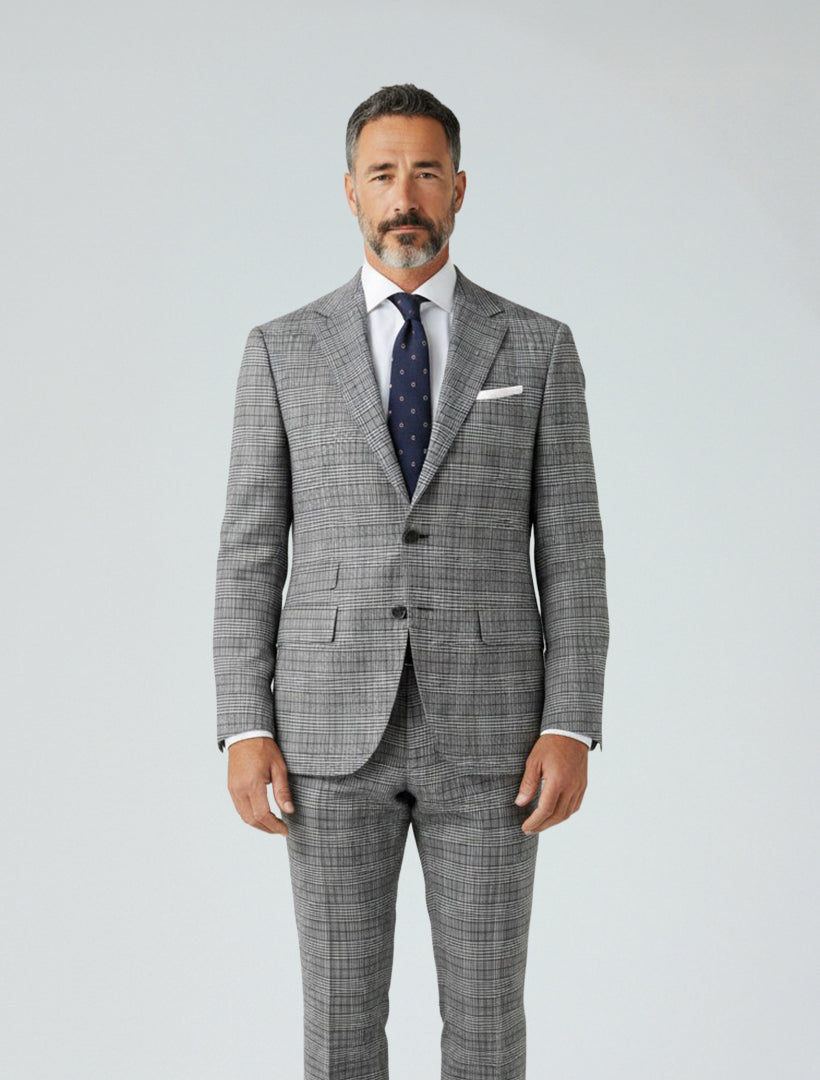 A man with short dark hair and a beard wears the Gianco Ferro Men's Light Grey Glen Plaid Slim Fit Suit, paired with a white shirt and navy patterned tie, standing against a plain light background.