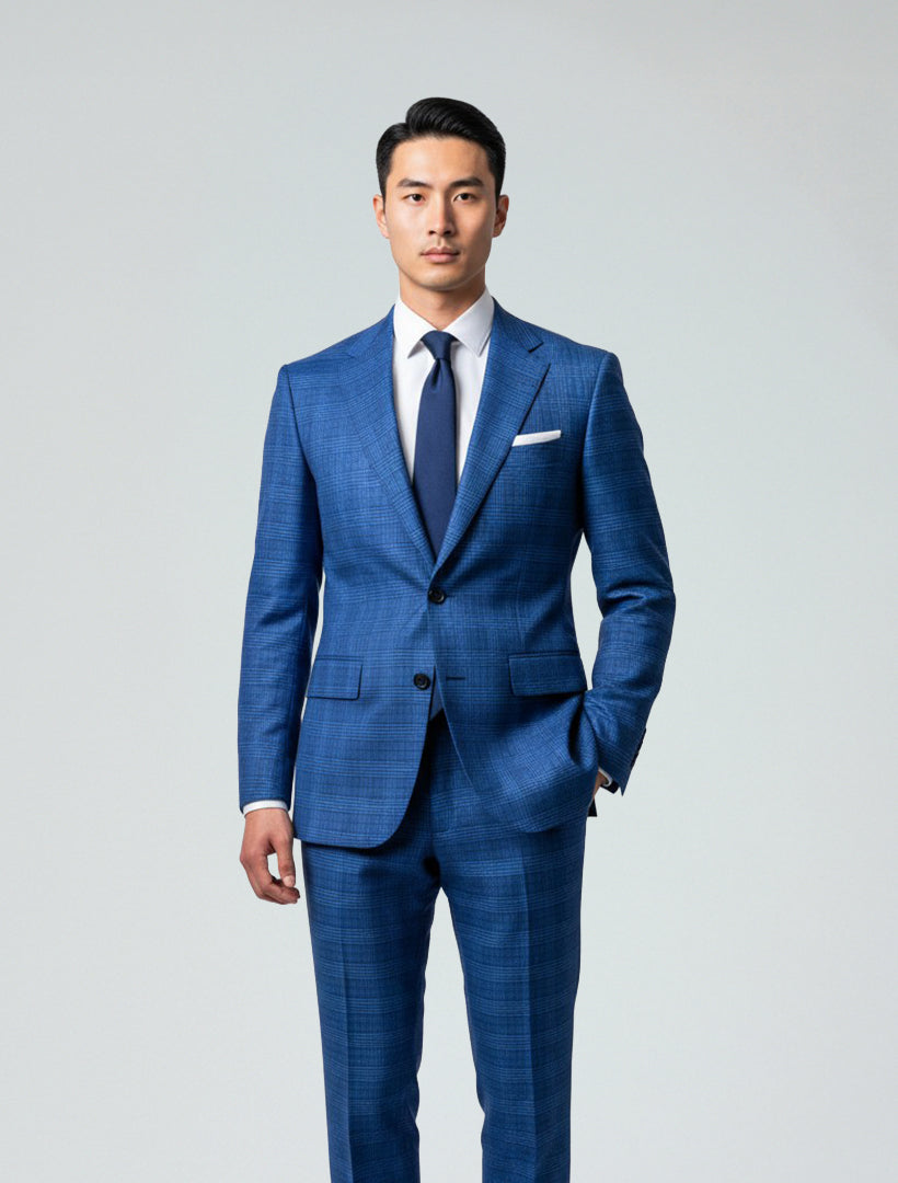 A man wears the Gianco Ferro Men's Blue Glen Plaid Slim Fit Suit with a white shirt, navy tie, and white pocket square, standing against a plain light background, facing forward with one hand in his pocket.