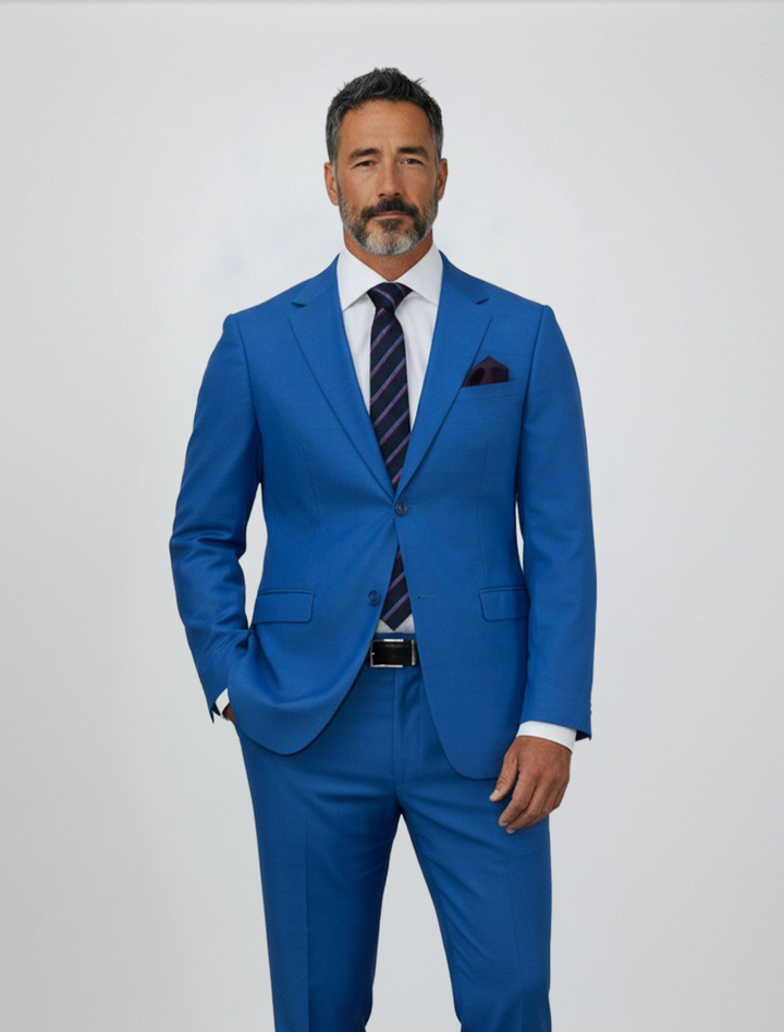 A man with short dark hair and a beard is seen wearing the Gianco Ferro Men’s Royal Blue Debossed Windowpane Slim Fit Suit, expertly crafted from premium Tetron Rayon, paired with a white shirt, striped tie, and pocket square against a plain light background.
