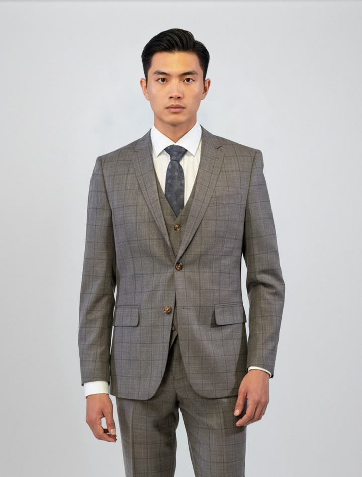 A man stands facing forward, wearing the FUBU Taupe with Brown Windowpane Vested Suit from Fubu The Colle, paired with a white shirt and dark patterned tie, against a plain light background.