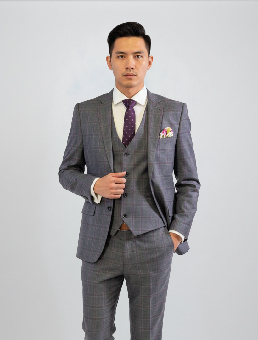 A man stands against a plain light background wearing the FUBU Charcoal with Purple Windowpane Vested Suit by Fubu The Colle, paired with a white shirt, purple polka dot tie, and patterned pocket square. The suit features a stretch armhole design for added comfort.