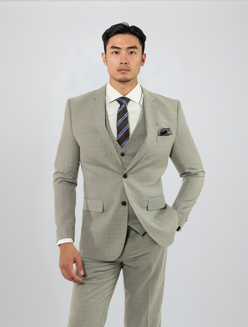 A man stands against a plain background wearing the FUBU Light Grey Solid Vested Suit from Fubu The Colle, featuring a modern fit with stretch armholes. He pairs it with a white dress shirt and striped tie, one hand in his pocket.