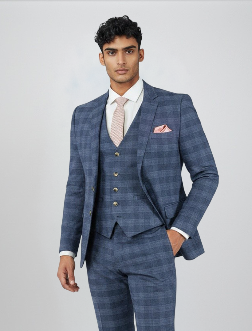 A man wearing the FUBU Sapphire Blue Glen Check Vested Suit by Fubu The Colle, styled with a white shirt, light pink tie, and matching pocket square, stands against a plain light background.