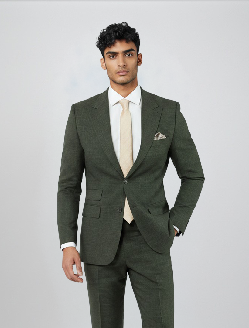 A man stands against a plain light background, wearing the FUBU Olive Green Peak Lapel 2-Piece Suit by Fubu The Colle. Crafted from premium Tetron Rayon in a modern slim fit, this suit features stretch armholes for added comfort and ease of movement. He pairs it with a white dress shirt, a light beige tie, and a matching pocket square, looking directly at the camera with one hand in his pocket.