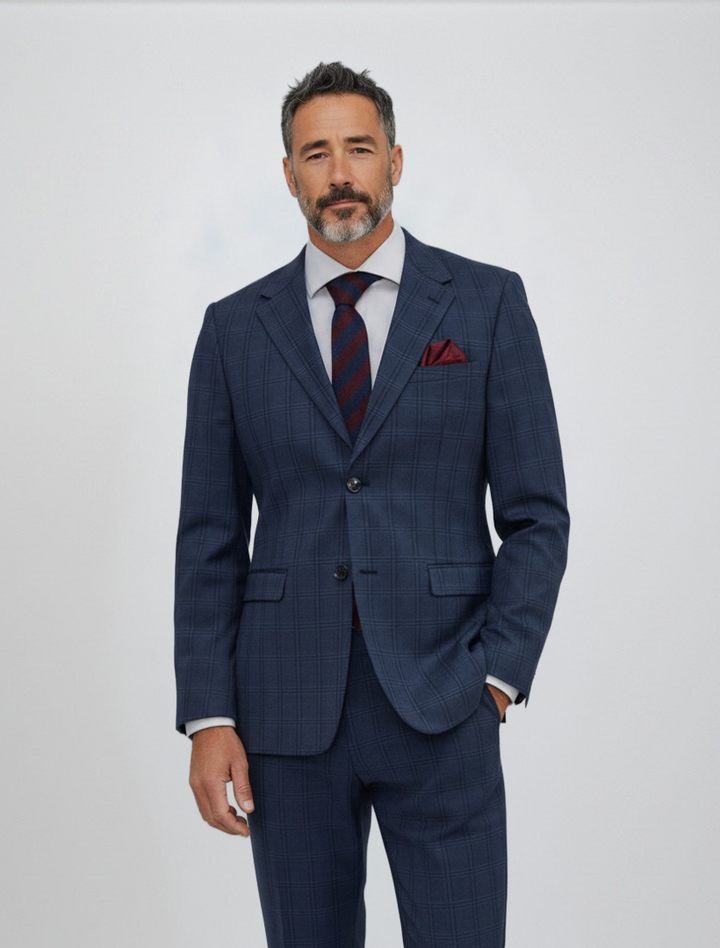 A man wears the Gianco Ferro Men’s Dark Blue with Black Double Windowpane Slim Fit Suit, paired with a white shirt, striped tie, and red pocket square, standing against a plain light background. Crafted from premium Tetron Rayon fabric and featuring a stretch armhole design, this suit blends style and comfort effortlessly.