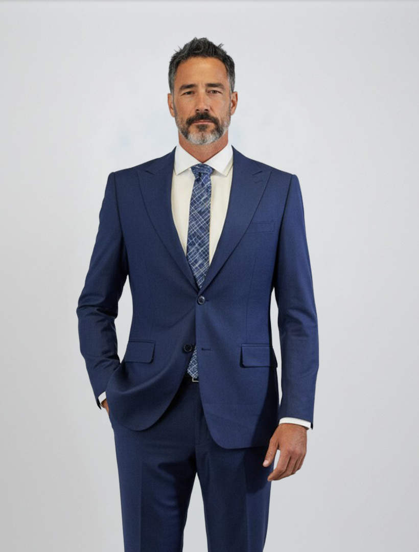 A man with short dark hair, a beard, and mustache is seen wearing the FUBU Indigo Solid Peak Lapel 2-Piece Suit by Fubu The Colle—a slim fit blue suit featuring a peak lapel jacket—paired with a white shirt and patterned tie, standing against a plain light background.
