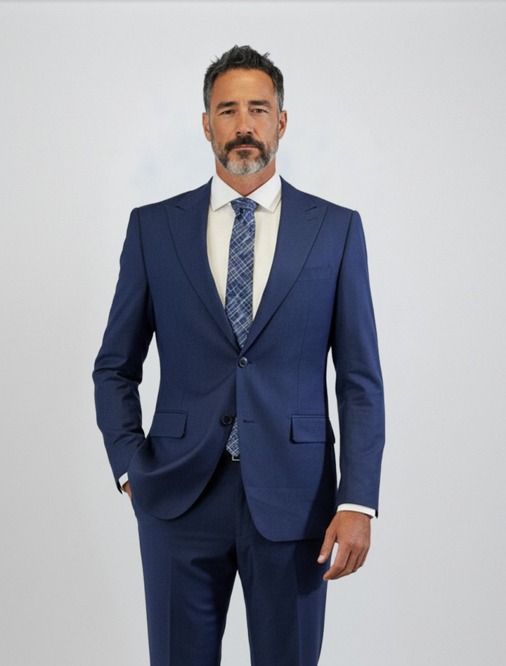 A man with short dark hair, a beard, and mustache is seen wearing the FUBU Indigo Solid Peak Lapel 2-Piece Suit by Fubu The Colle—a slim fit blue suit featuring a peak lapel jacket—paired with a white shirt and patterned tie, standing against a plain light background.