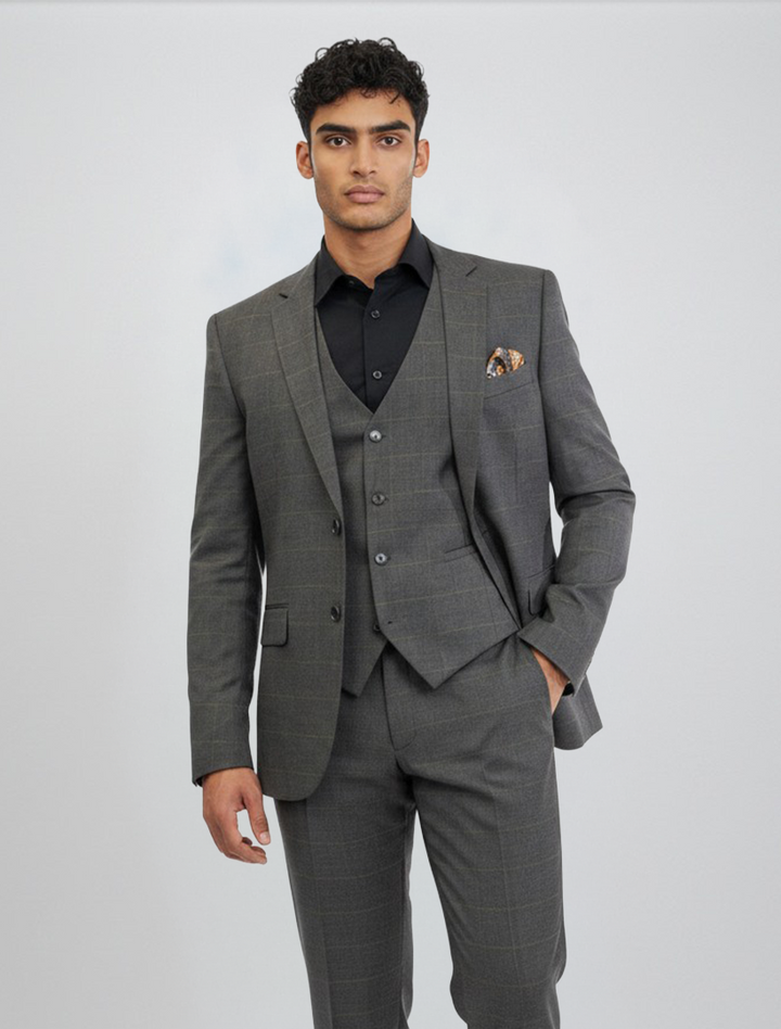 A man wearing the FUBU Charcoal Grey Windowpane Vested Suit by Fubu The Colle, featuring a slim fit and stretch armhole design for extra comfort, paired with a black shirt and standing against a plain light background with one hand in his pocket.