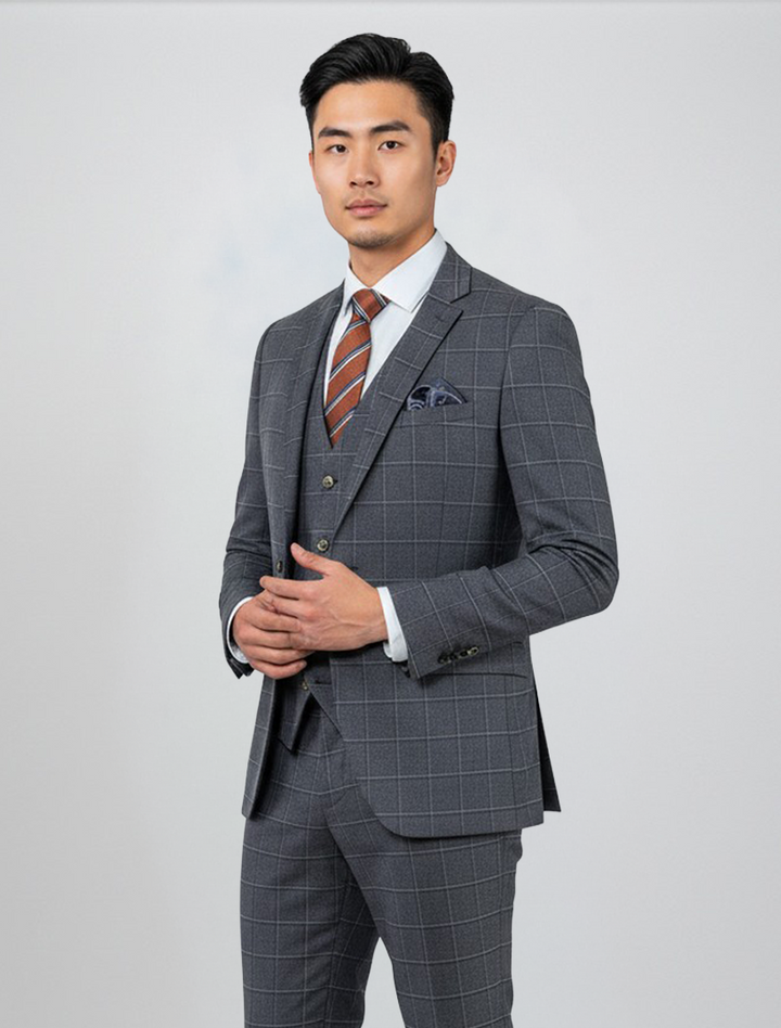 A man stands against a plain background wearing the Fubu The Colle FUBU Navy Pin Check Windowpane Vested Suit, paired with a white shirt and striped brown tie, his hands lightly clasped in front of him.