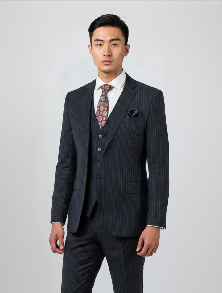 A man stands facing forward against a plain light background, wearing the FUBU Navy with White Pinstripe Vested Suit by Fubu The Colle, paired with a white shirt, patterned tie, and pocket square.