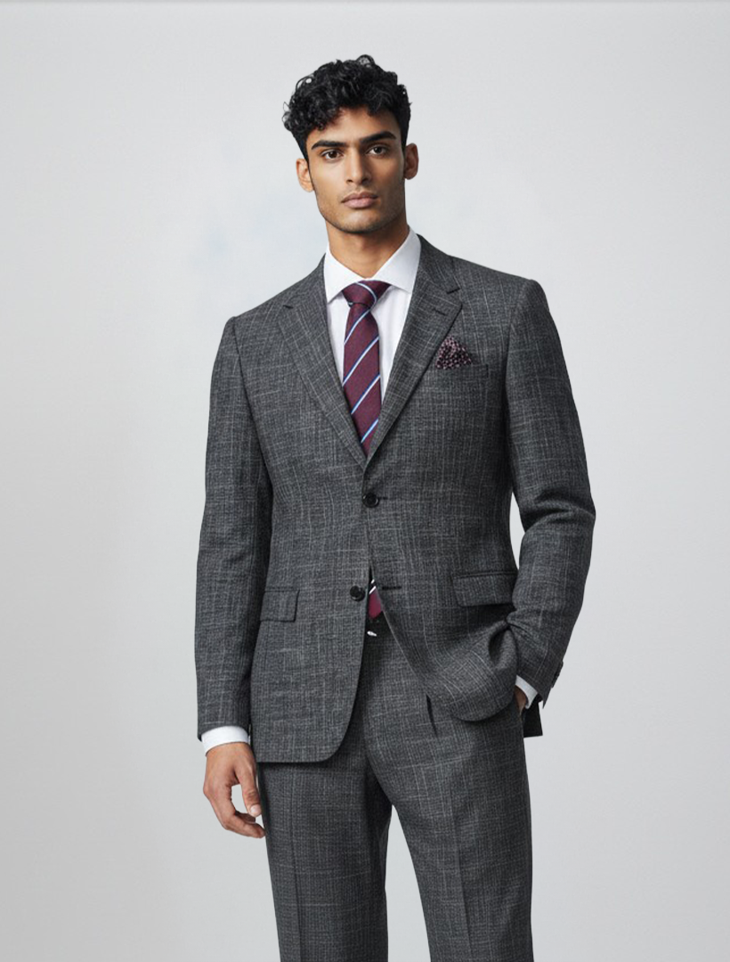 A man wearing the Gianco Ferro Men’s Charcoal with Black Pattern Slim Fit Suit, expertly crafted from premium Tetron Rayon, stands against a plain light background with one hand in his pocket. The suit is paired with a white shirt, striped tie, and pocket square, and features a stretch armhole design for enhanced comfort and mobility.