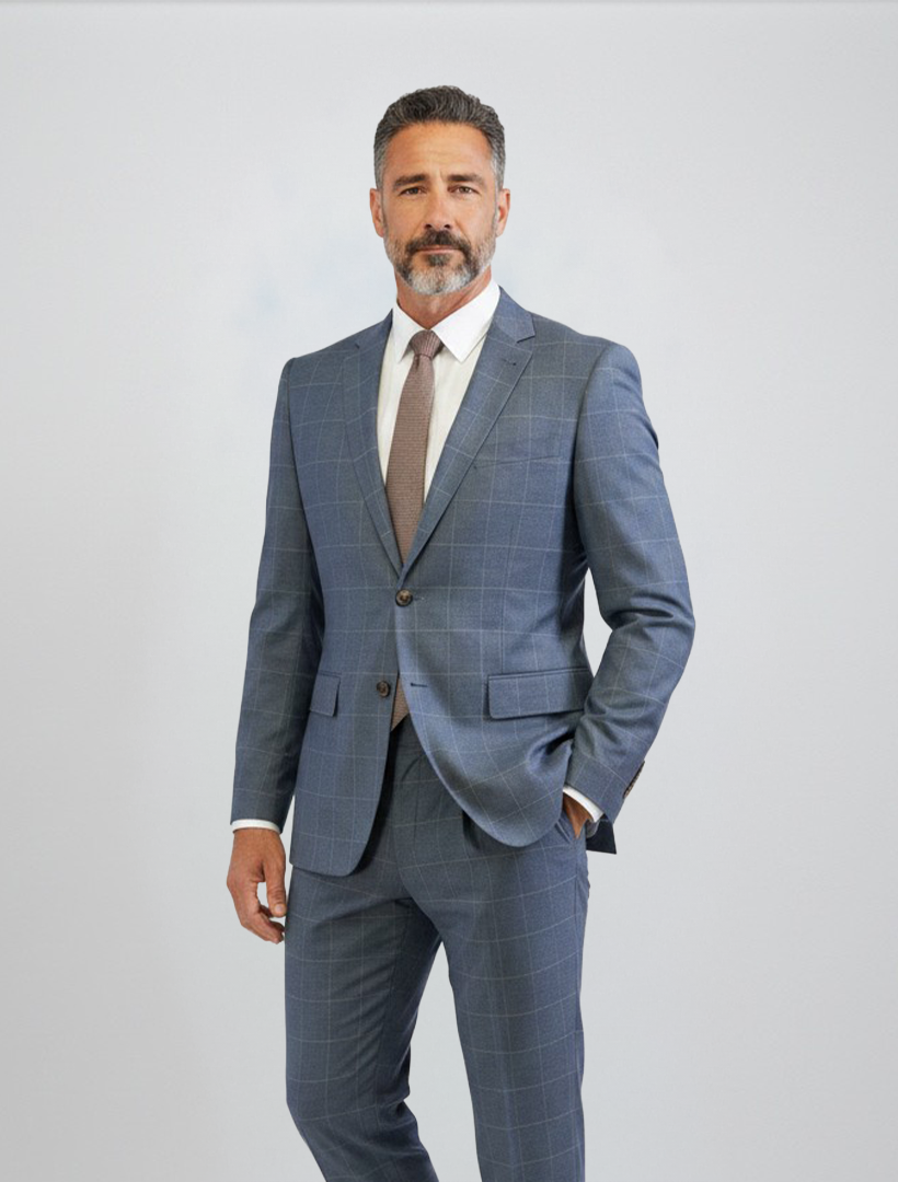 A man with short gray hair and a beard wears the FUBU Gravel Blue with Peach Windowpane 2-Piece Suit by Fubu The Colle, featuring a slim fit crafted from Tetron Rayon. Paired with a white shirt and brown tie, he stands against a plain light background with one hand in his pocket, while the suit’s stretch armhole provides both comfort and style.